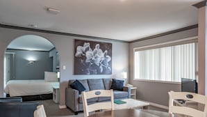 A hotel room with a bed, a gray sofa, a dining table, and a large window with blinds. - Tiki Shores Inn & Suites (Penticton)