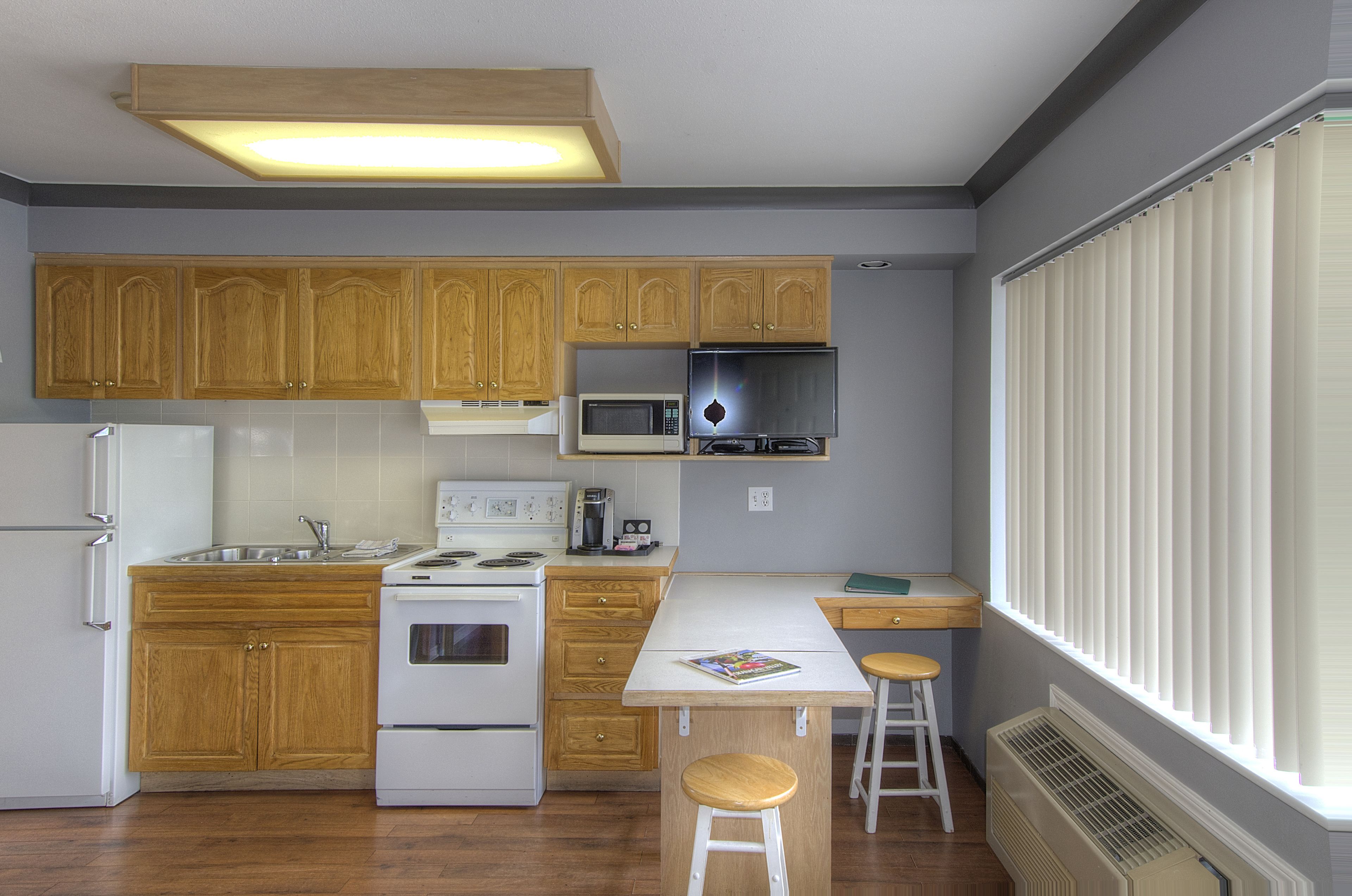 A compact kitchen with wooden cabinets, a microwave, stove, and a window with blinds.