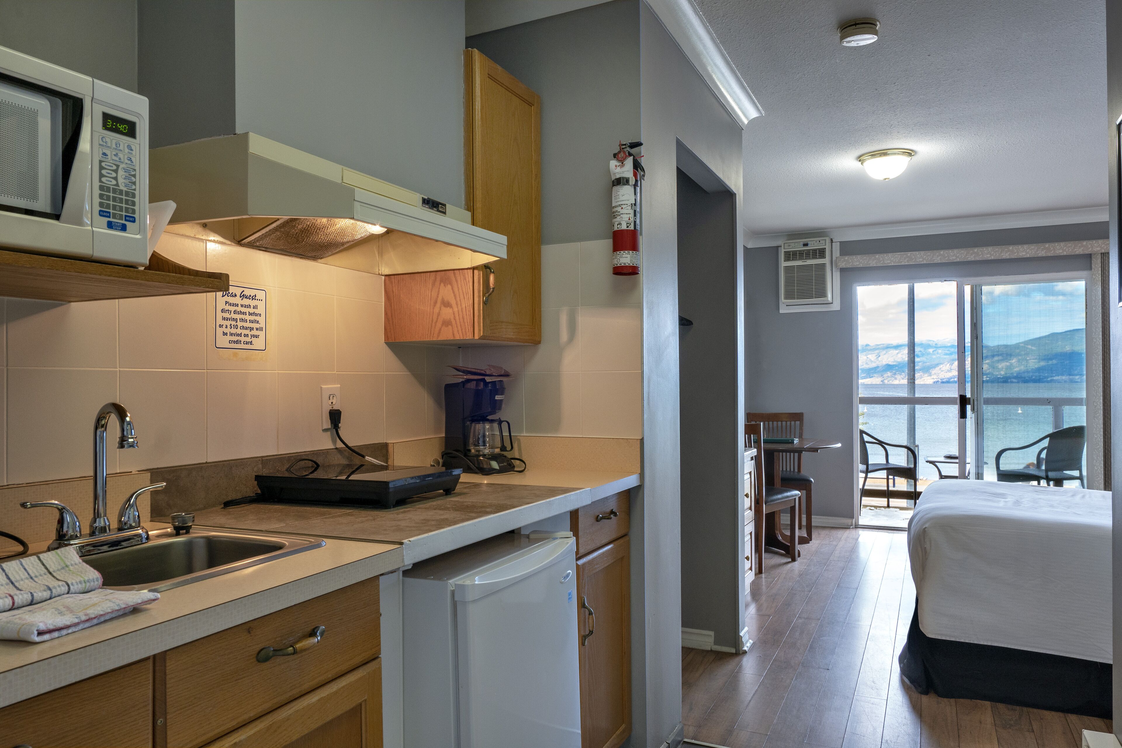 A modern kitchen with a microwave, sink, and coffee maker. There is a sign on the wall, a fire extinguisher, and a view of the balcony with chairs.