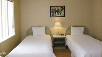 Una habitación de hotel con dos camas individuales, una mesita de noche con una lámpara, un cuadro de uvas en la pared y una ventana con persianas.