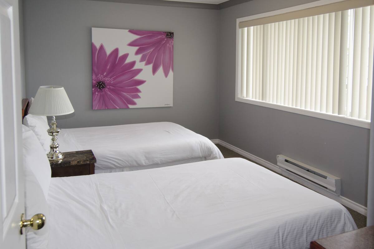 A bedroom with two beds, a nightstand, a lamp, a window with blinds, and a wall-mounted air conditioner.