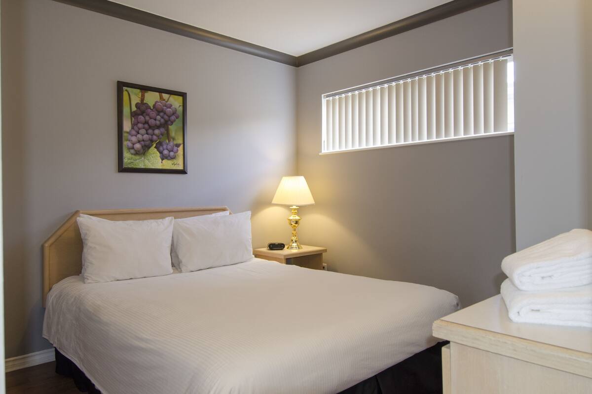 A neatly made bed with white linens, a bedside lamp, and a framed picture of grapes on the wall.