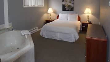 A hotel room with a large bed, two bedside tables with lamps, a wooden nightstand, a white bathtub, and a framed floral artwork on the wall.