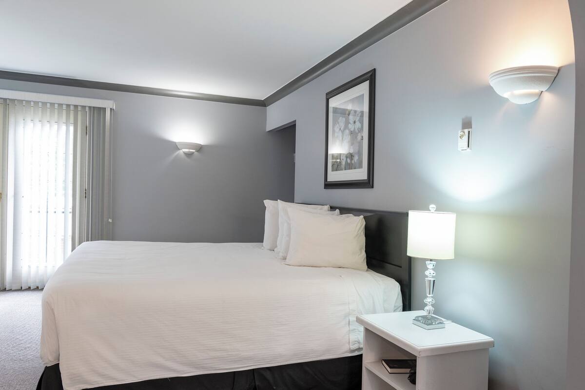 A hotel room with a large bed, a bedside table with a lamp, a framed picture on the wall, and a window with blinds.