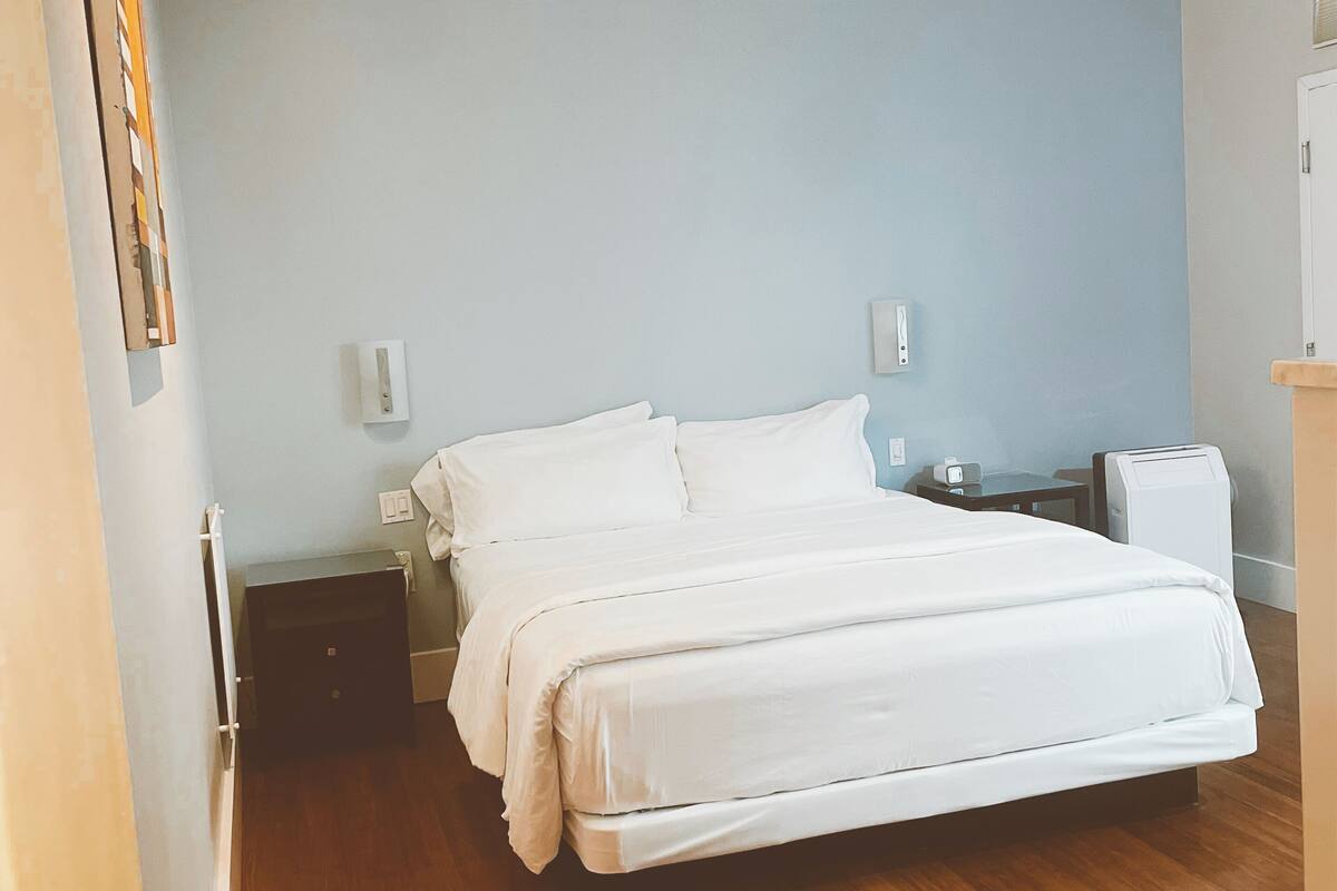 A bedroom with a large bed, wooden flooring, a ceiling fan, and a nightstand with a lamp.