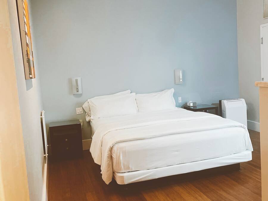 A bedroom with a large bed, wooden flooring, a ceiling fan, and a nightstand with a lamp.