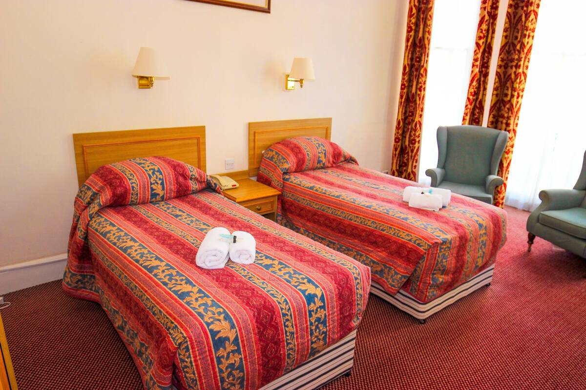A hotel room with two beds, each with patterned bedding and towels, a green armchair, and a lamp on the wall.