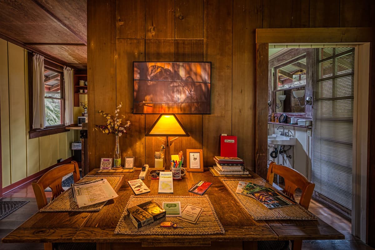 A wooden dining table with placemats, a vase with flowers, a lamp, and various items including books and a red box.