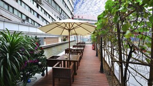 A covered outdoor seating area with wooden tables and a large umbrella, surrounded by greenery and a view of a building with a tiled facade. - Sunon Hotel (Shenzhen)