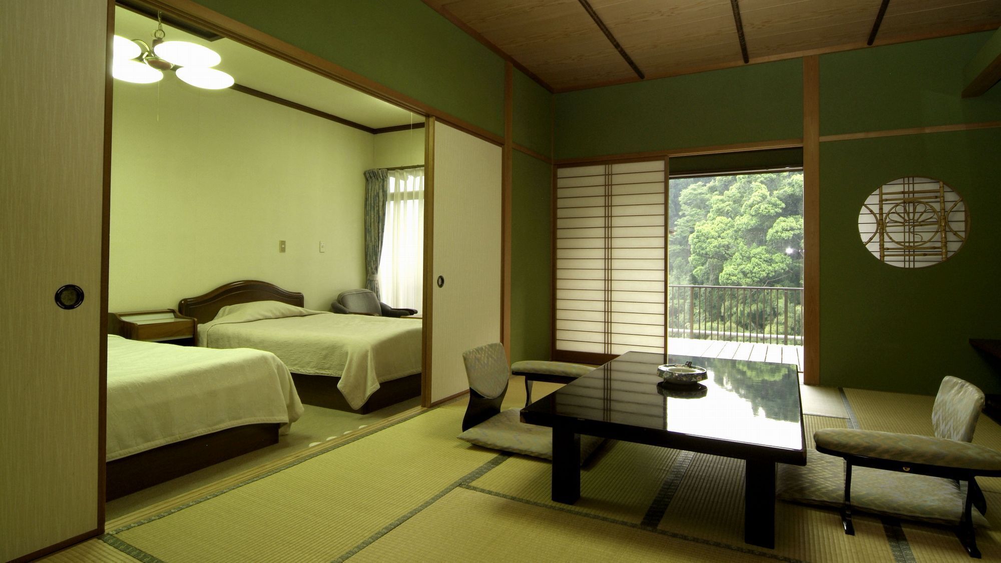 A traditional Japanese-style room with two beds, a tatami mat floor, a low table with a chair, and a view of greenery outside.