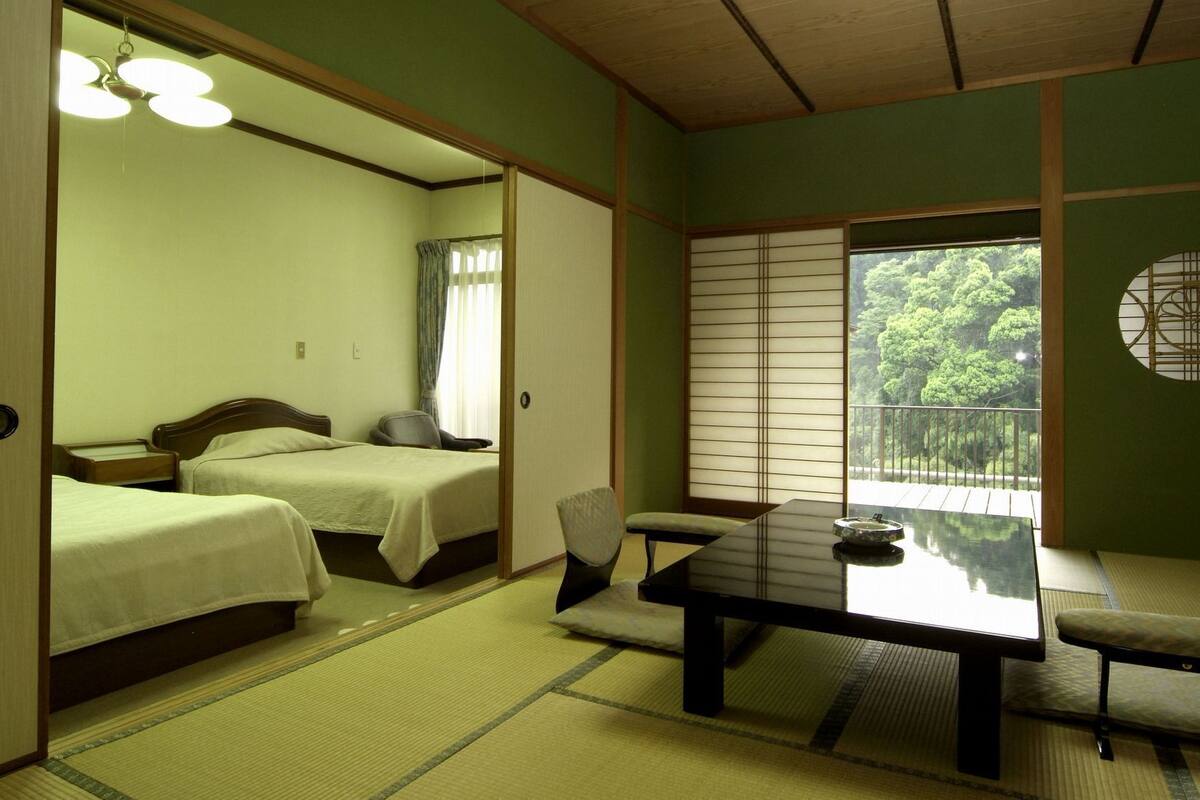 A traditional Japanese-style room with two beds, a tatami mat floor, a low table with a chair, and a view of greenery outside.