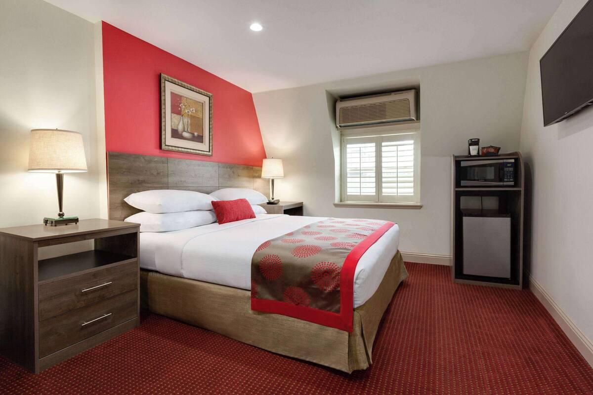 A hotel room with a bed, two bedside tables, a TV, and a window with blinds.