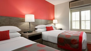 Habitación de hotel con dos camas, cada una con una colcha roja y blanca, cabeceros de madera y una ventana con persianas.