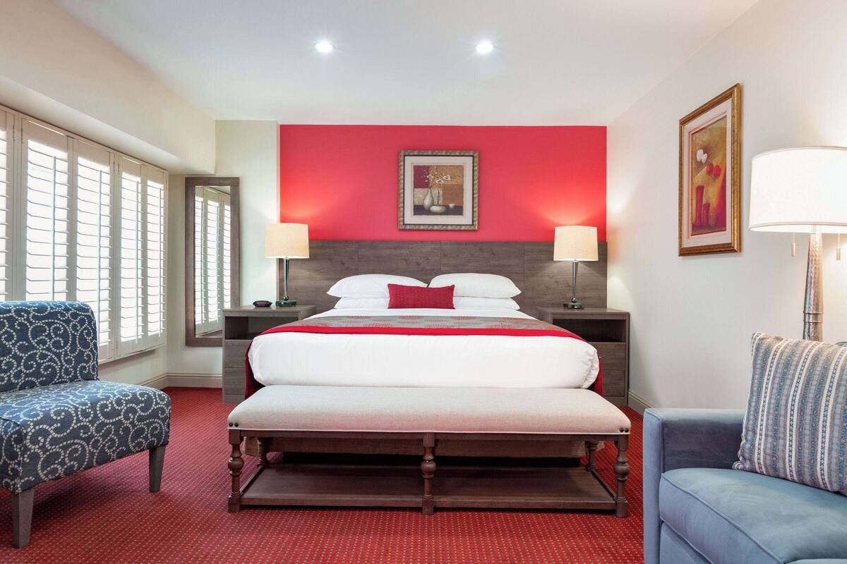 A hotel room with a large bed, two bedside tables with lamps, a blue armchair, a red carpet, and a window with blinds.