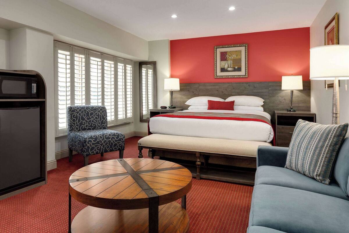 A hotel room with a bed, a sofa, a round wooden table, a chair, a TV, and a window with blinds.