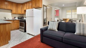 Un espacio habitable compacto con área de cocina que incluye gabinetes de madera, refrigerador, microondas y estufa. Hay un sofá de color oscuro con cojines estampados y una alfombra roja en el suelo.