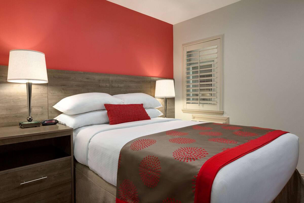 A hotel room with a bed, two bedside lamps, a nightstand, and a window with blinds.