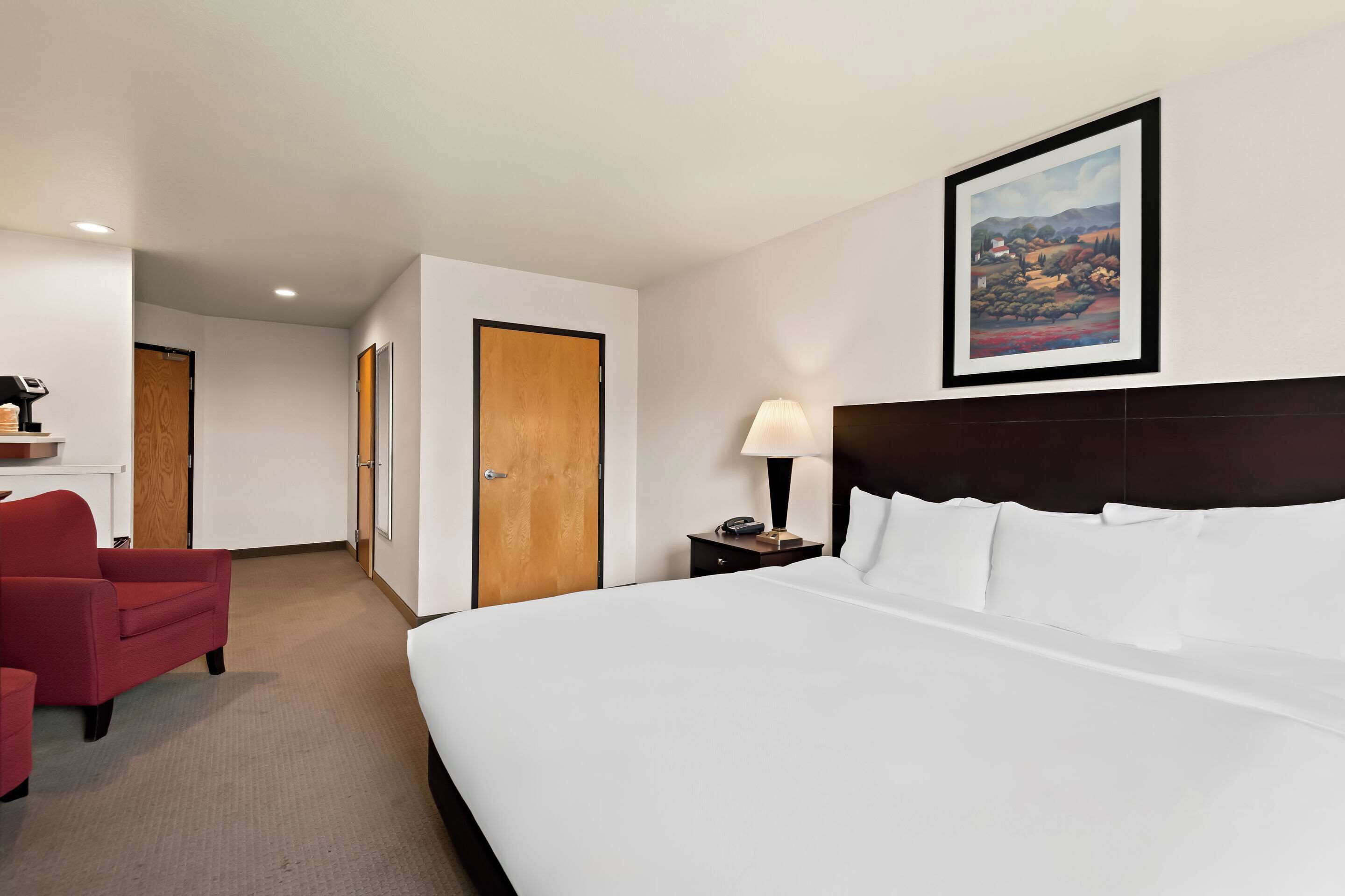 A hotel room with a large bed, a red armchair, a nightstand with a lamp, a framed painting on the wall, and a wooden door.