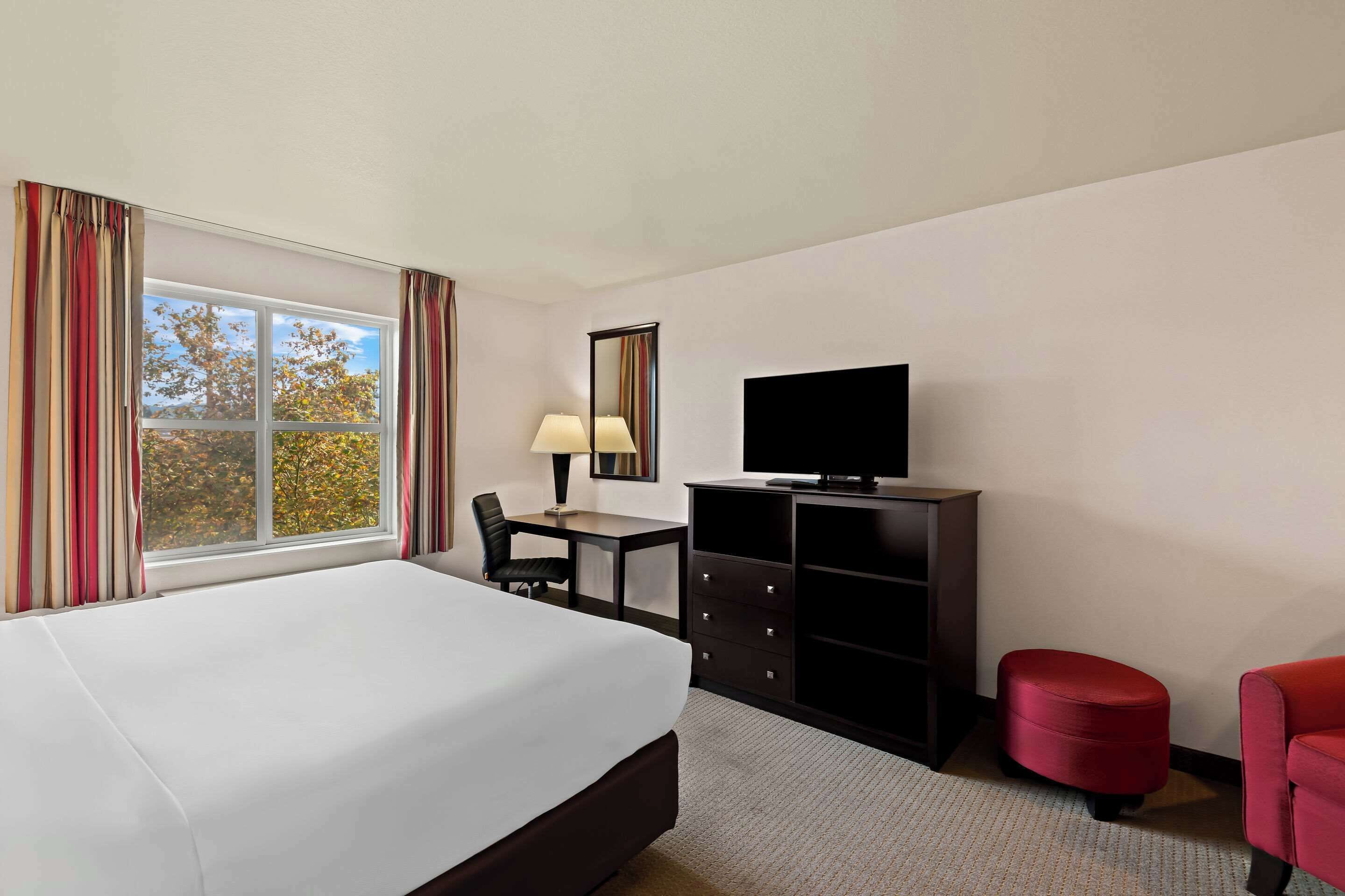 A hotel room with a large bed, a desk with a chair, a television, a red ottoman, and a window with curtains.