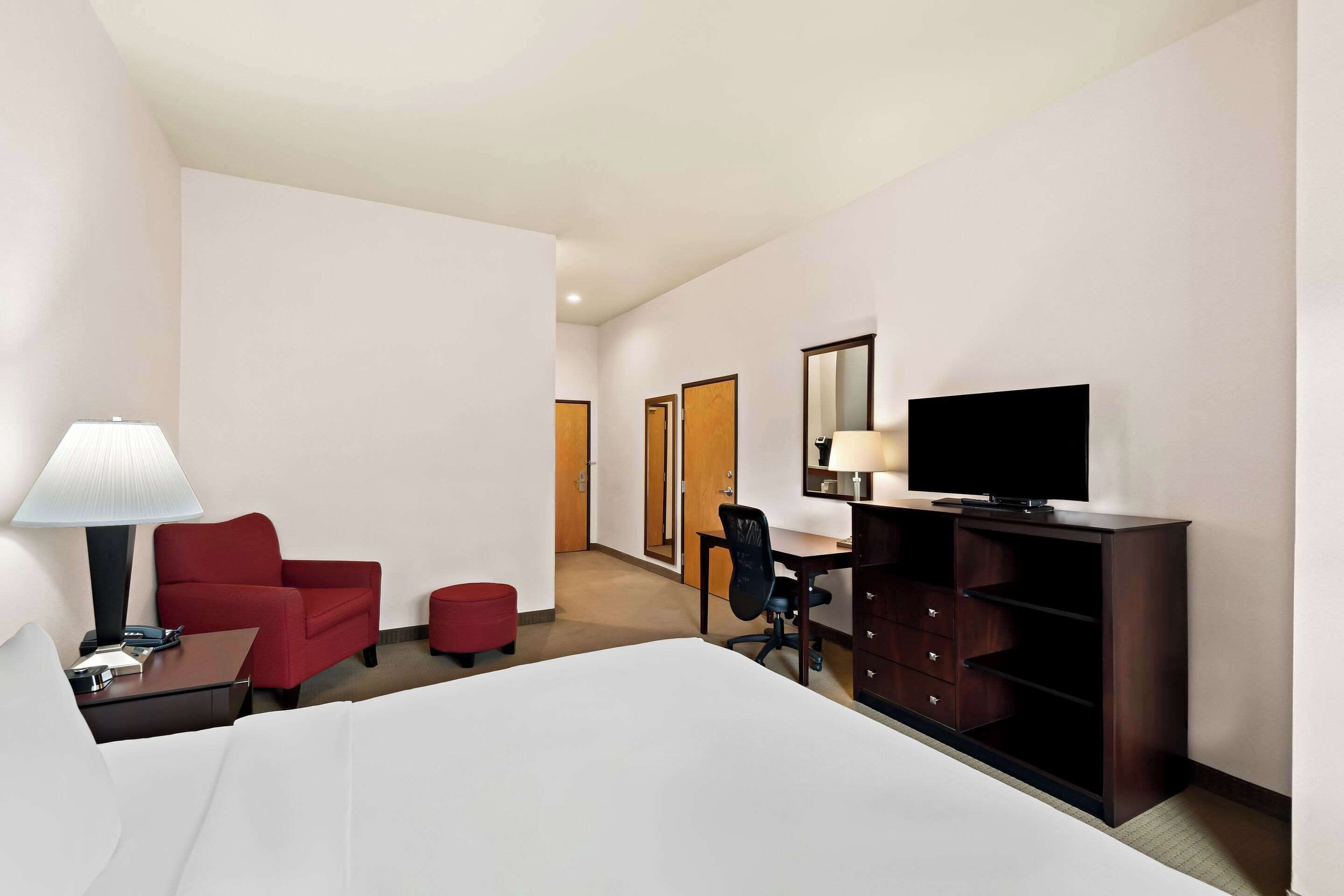 A hotel room with a bed, a desk with a chair, a TV, a lamp, and a red chair.