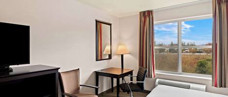 Habitación de hotel con cama, escritorio, silla, televisor, lámpara y ventana con cortinas.