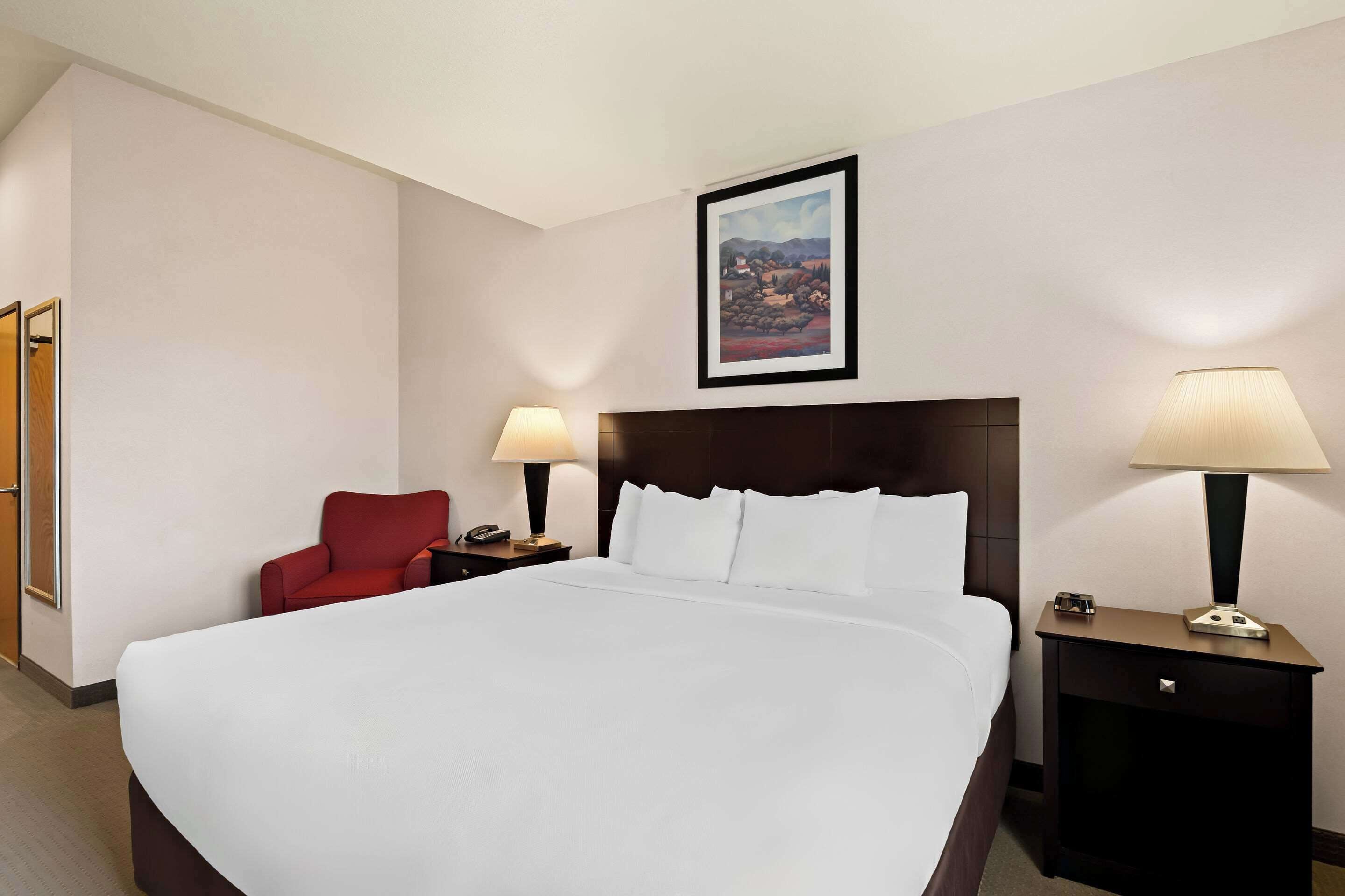 Une chambre d’hôtel avec un grand lit, un fauteuil rouge, deux tables de chevet avec des lampes et un tableau encadré au mur.