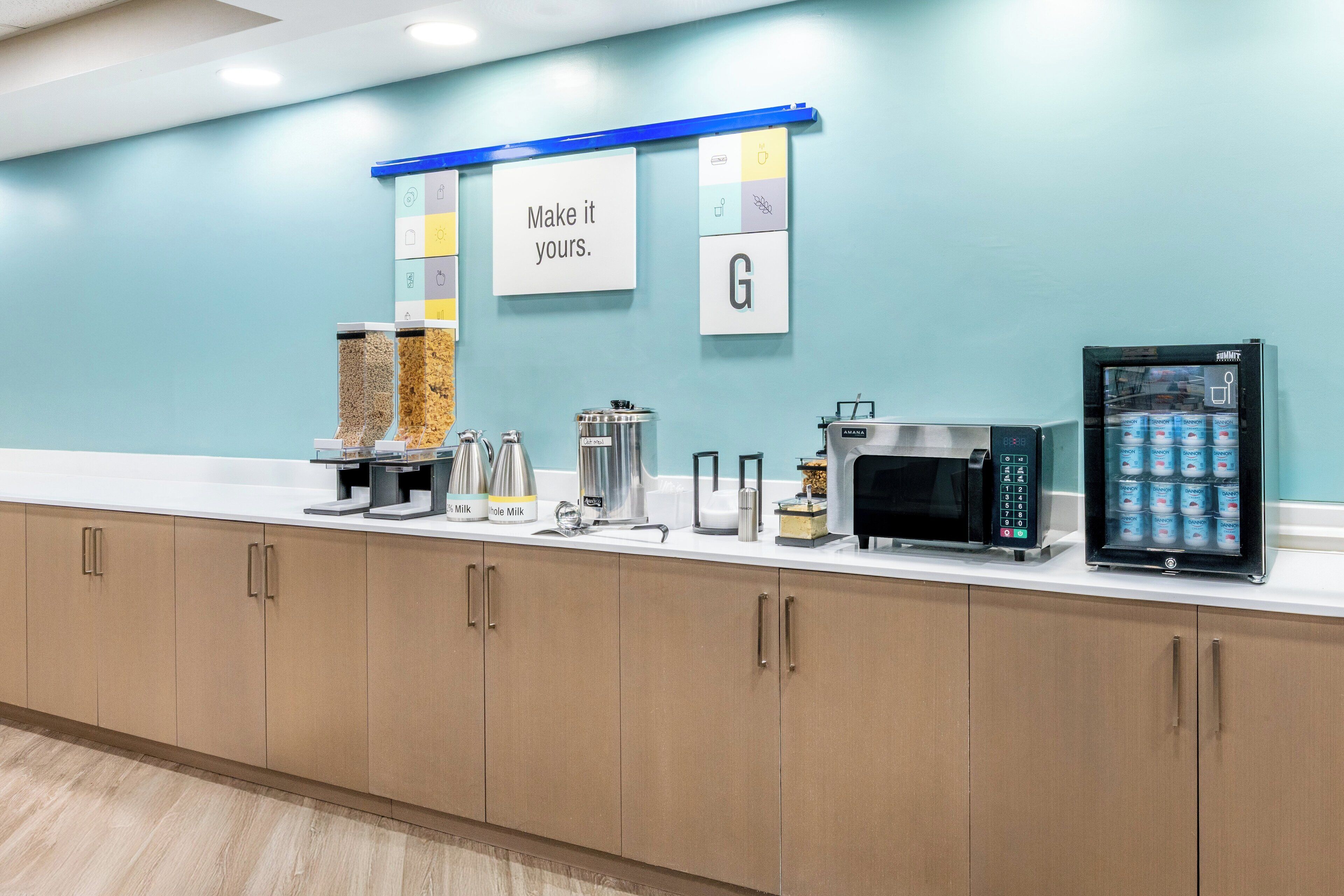 Un área de cocina moderna con pared azul, máquina de café, microondas, refrigerador y un letrero que dice “Hazlo tuyo”.