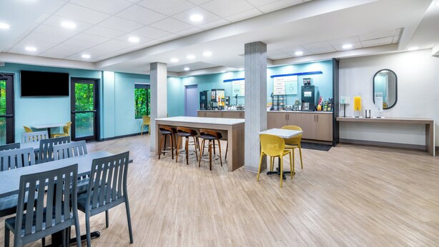 A spacious indoor area with a long table and chairs, a counter with a coffee machine, and a large mirror on the wall. - Garner Hotel Doswell Southeast by IHG (Doswell)