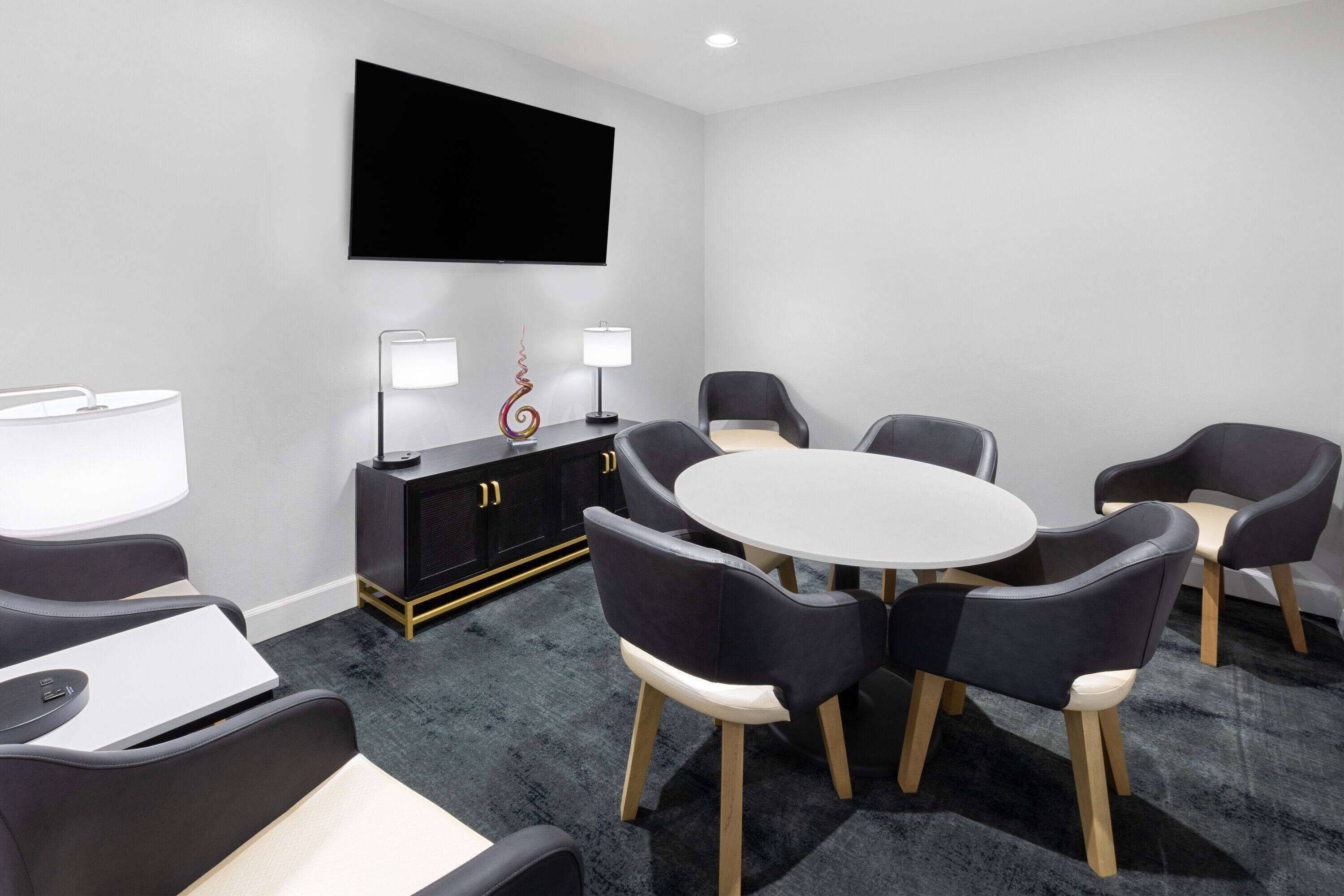 Une salle de réunion moderne équipée d’une table ronde, de six chaises, d’un téléviseur à écran plat, d’un meuble noir avec deux lampes et d’un sol recouvert de moquette.