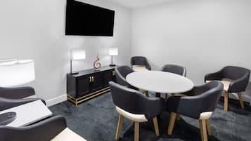 A modern meeting room with a round table, six chairs, a flat-screen TV, a black cabinet with two lamps, and a carpeted floor.