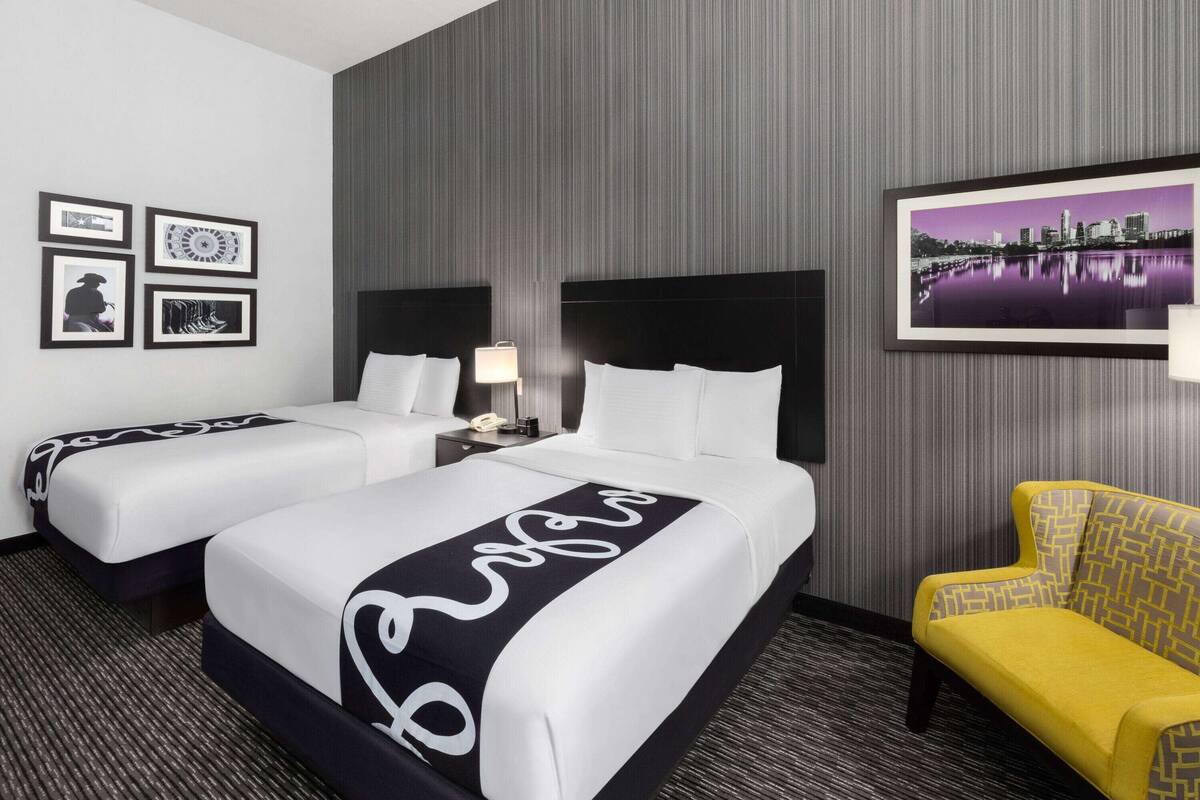 Una habitación de hotel con dos camas, una mesita de noche, una lámpara, una silla y cuadros en la pared.