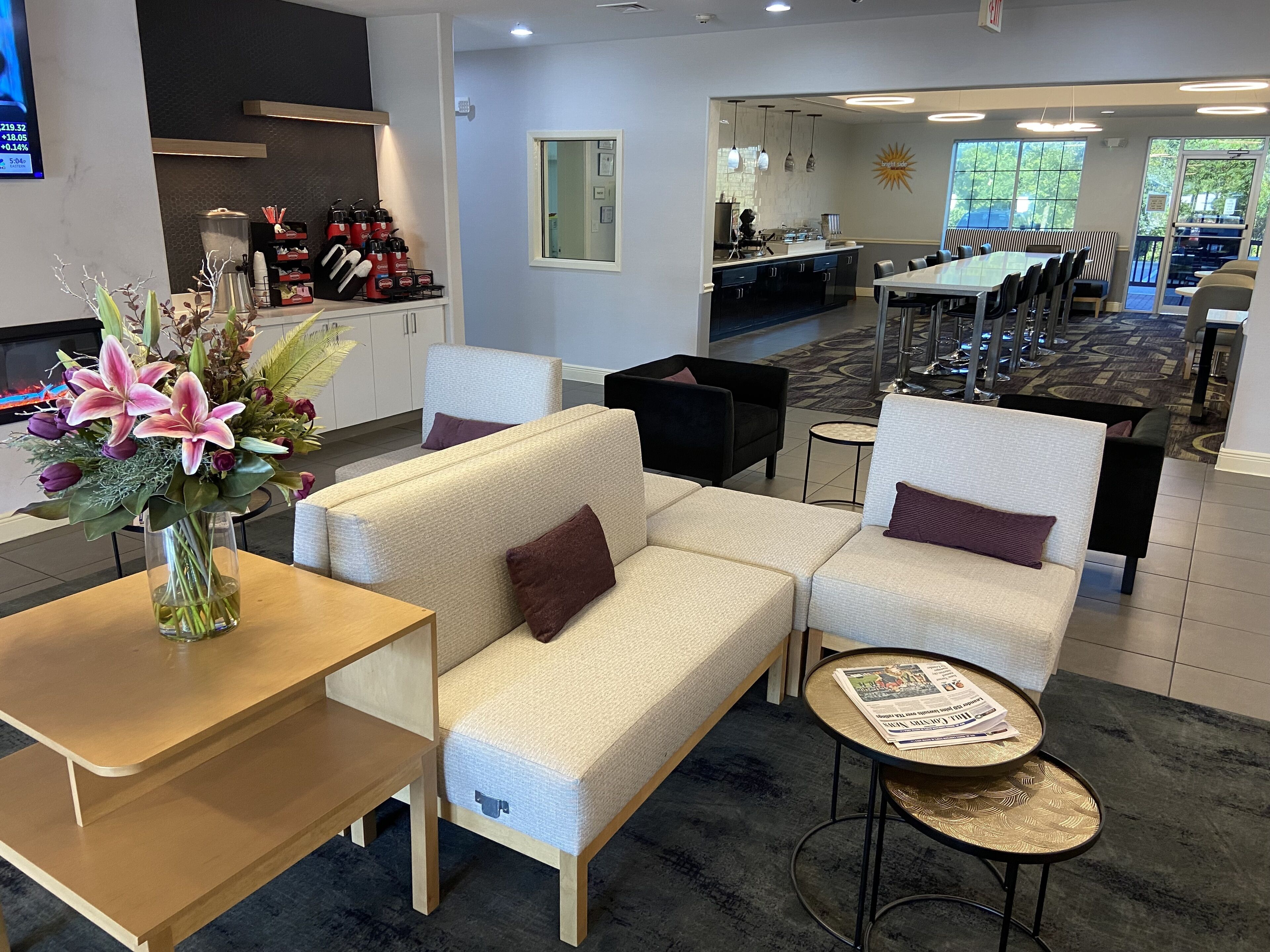A hotel lobby with a sofa, chairs, a small table, a vase of flowers, and a kitchen area in the background.