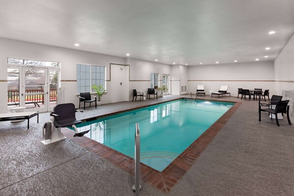A large indoor swimming pool with a tiled deck, surrounded by black chairs and tables, and a view of a fenced outdoor area through glass doors.