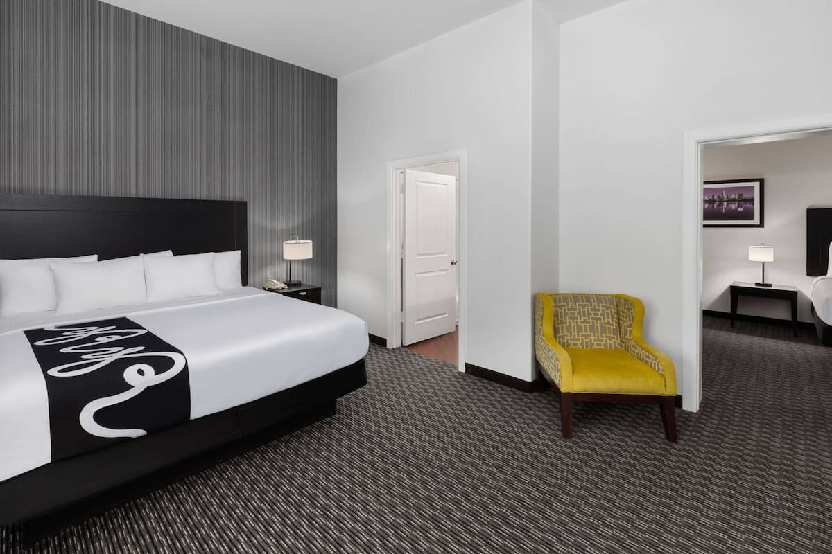 Habitación de hotel con una cama grande, una mesita de noche, una lámpara, una silla y una puerta que da a otra habitación.