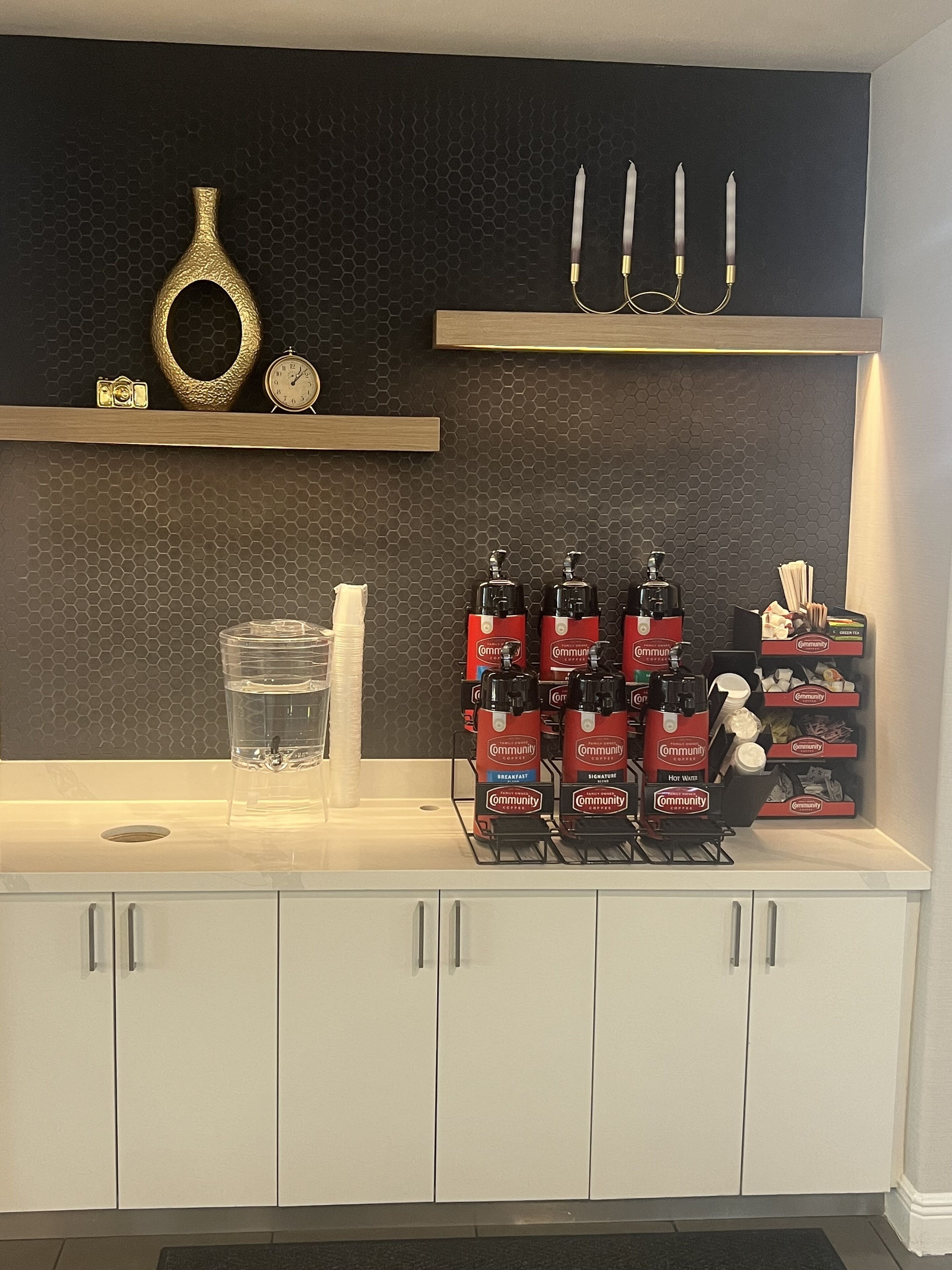 Una zona de cocina con un salpicadero hexagonal negro, una encimera blanca y un mueble bajo de color blanco. Hay tres dispensadores rojos con la etiqueta “Community Coffee” sobre la encimera, un tarro de cristal transparente y una toalla blanca colgada en la pared.