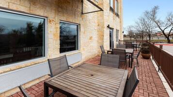 An outdoor patio with brick flooring, wooden tables and chairs, and large windows.