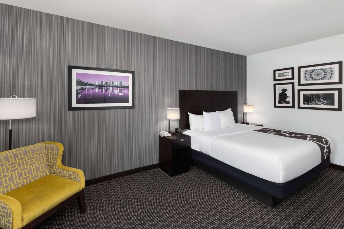 Habitación de hotel con cama, mesitas de noche, lámpara, una silla e imagen enmarcada en la pared.