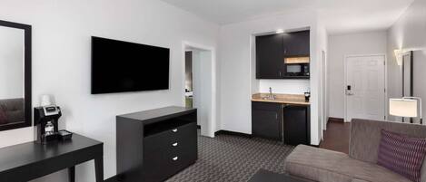 Habitación de hotel compacta con kitchenette, televisor de pantalla plana, sofá, máquina de café y escritorio.