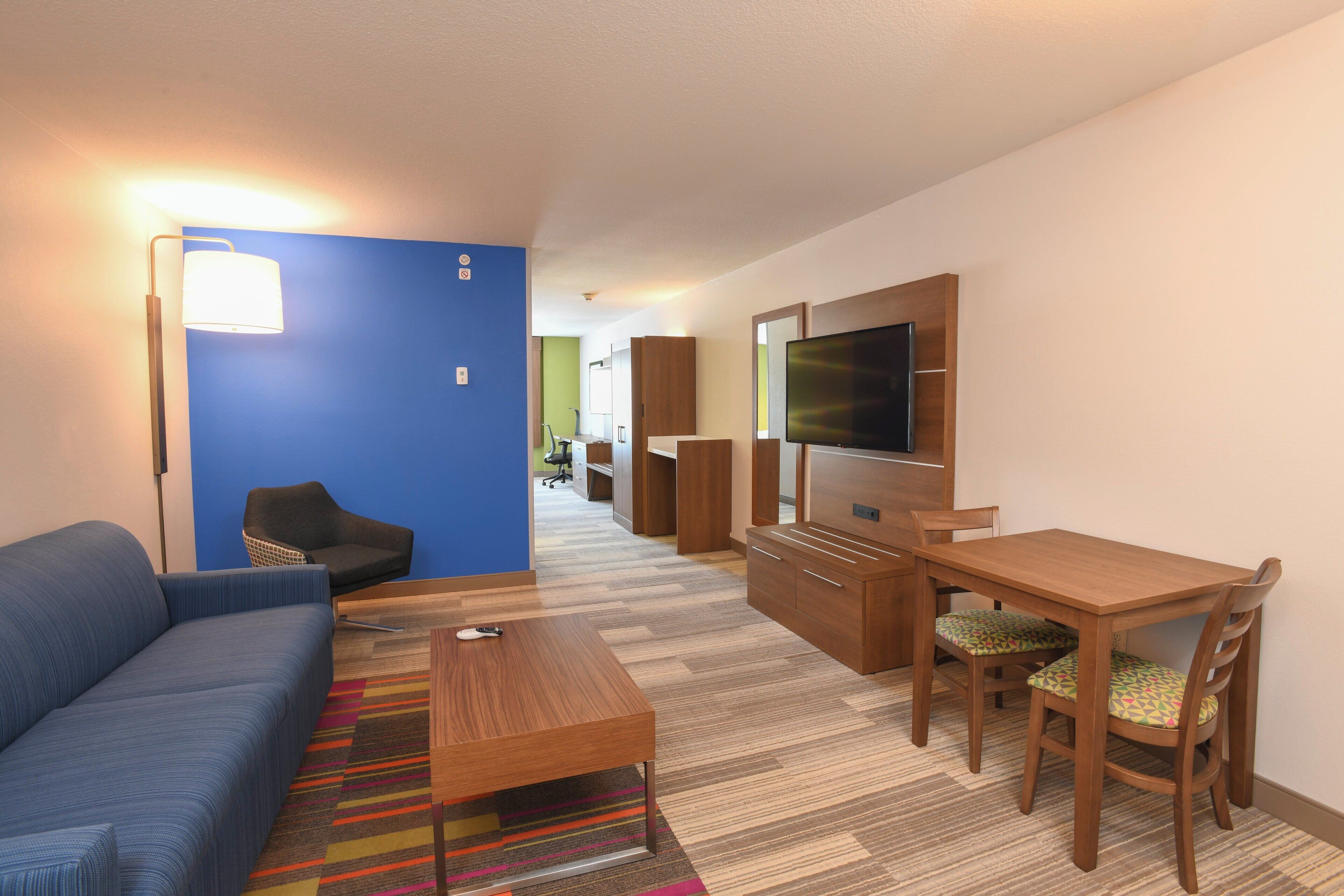 Habitación de hotel moderna con una pared azul de acento, un sofá, un escritorio de madera, un televisor y una alfombra colorida a rayas.