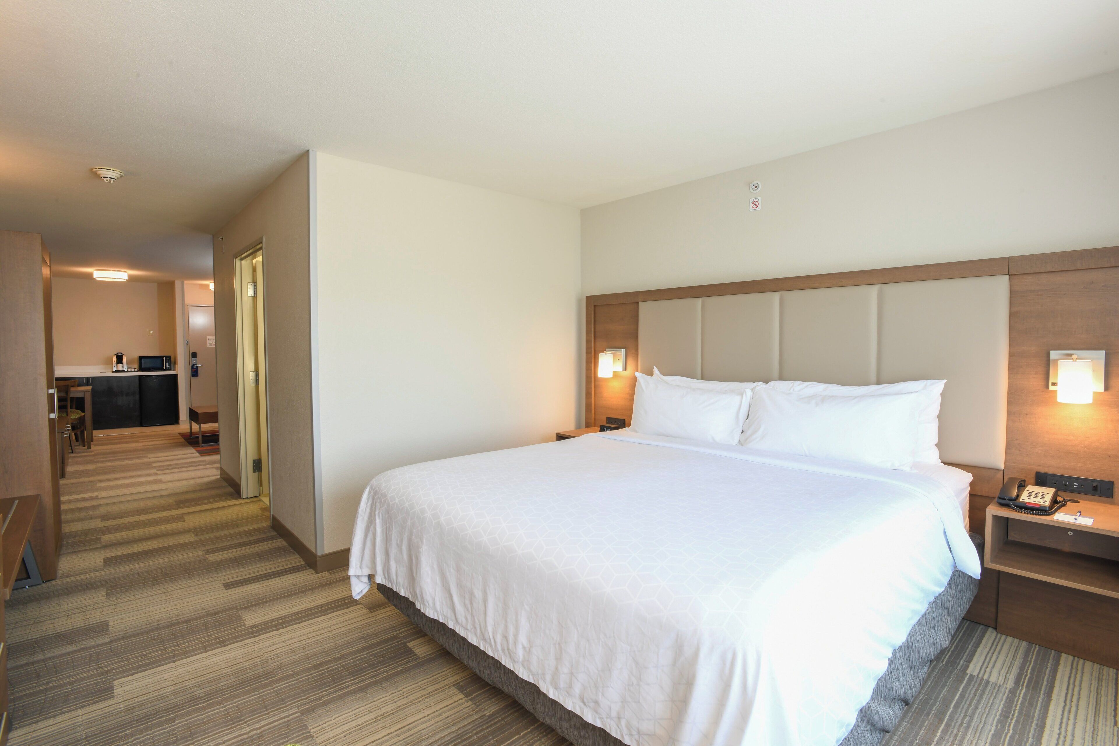 A hotel room with a large bed, a bedside table with a phone, a nightstand with a lamp, and a kitchenette area in the background.