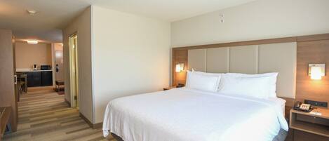 A hotel room with a large bed, a bedside table with a phone, a nightstand with a lamp, and a kitchenette area in the background.
