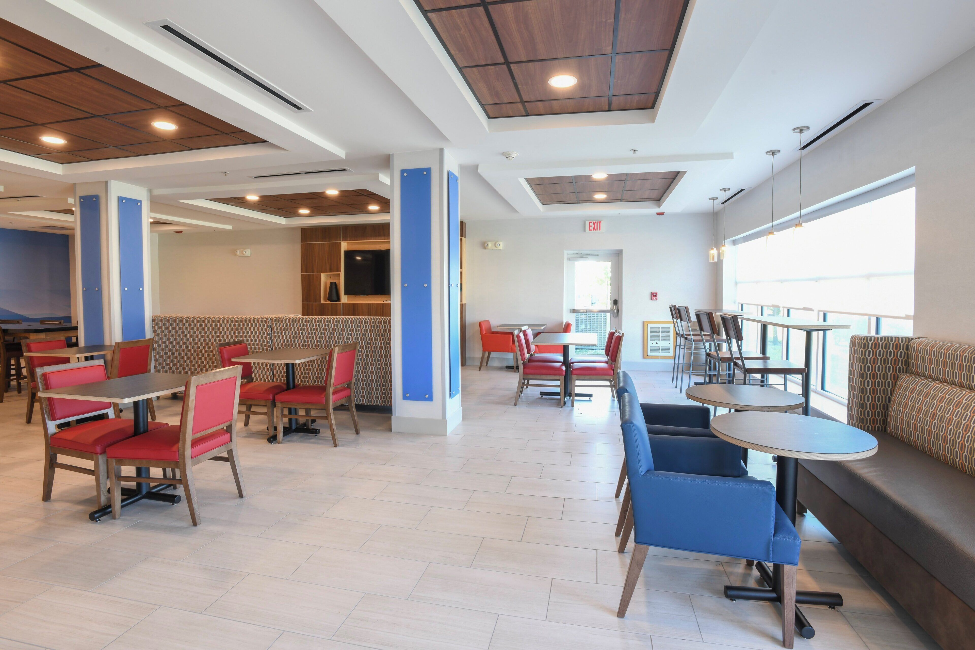 A spacious indoor dining area with multiple seating options including booths and tables, a visible exit sign, and a large window providing natural light.