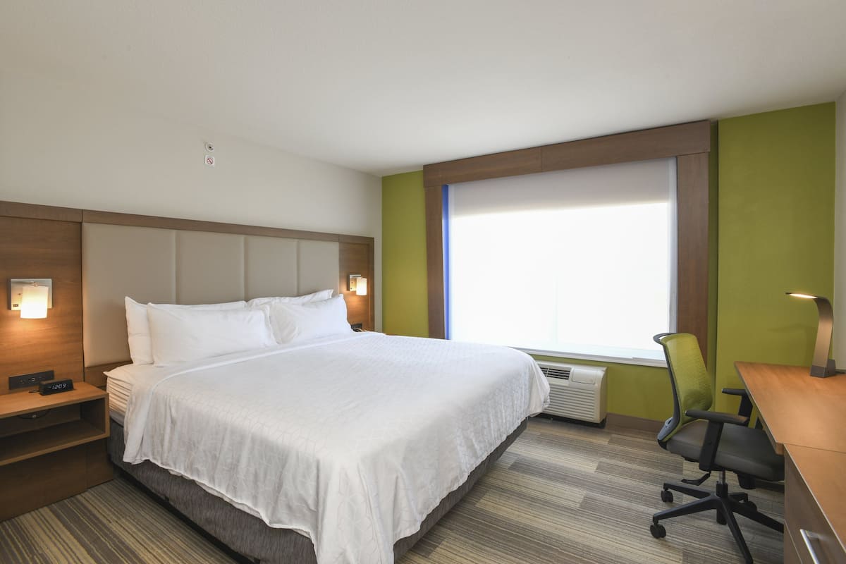 Une chambre d’hôtel avec un grand lit, un bureau, une chaise, une table de chevet et une fenêtre avec des stores.