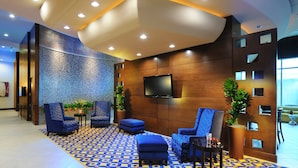 A lobby with a patterned floor, seating area with blue upholstered chairs, a flat-screen TV mounted on the wall, and potted plants. - Houston Marriott Energy Corridor (Houston)