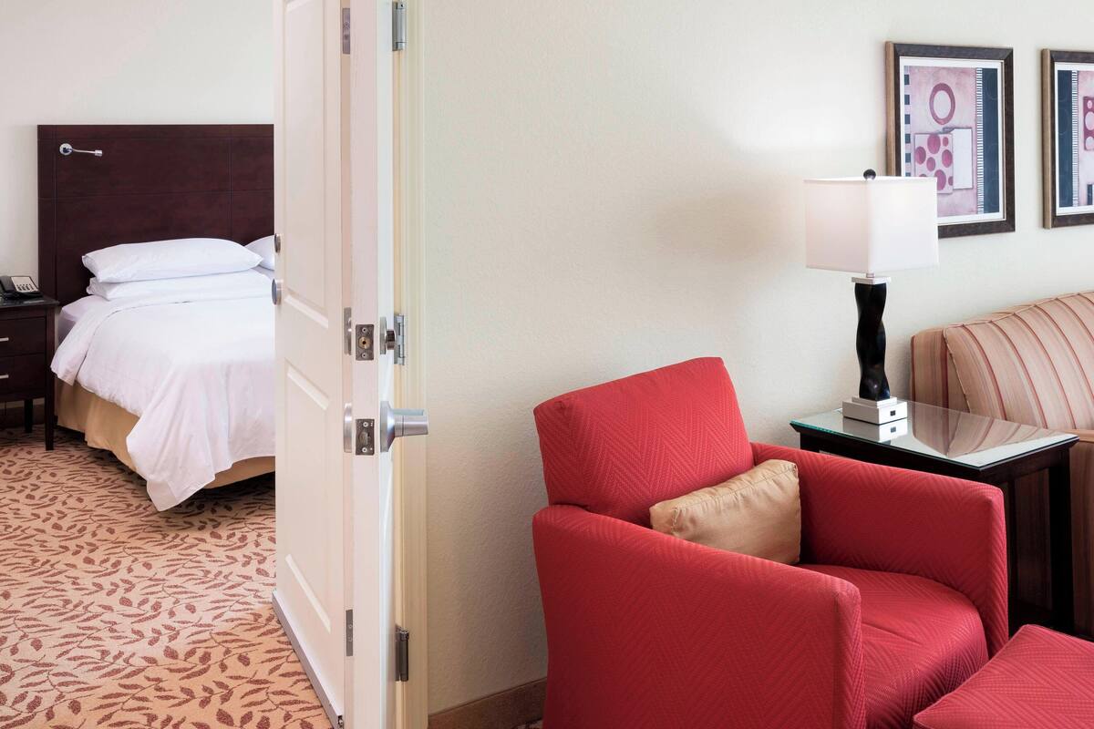 Una camera d'albergo con un letto, una poltrona rossa, un comodino, una lampada e quadri appesi al muro.