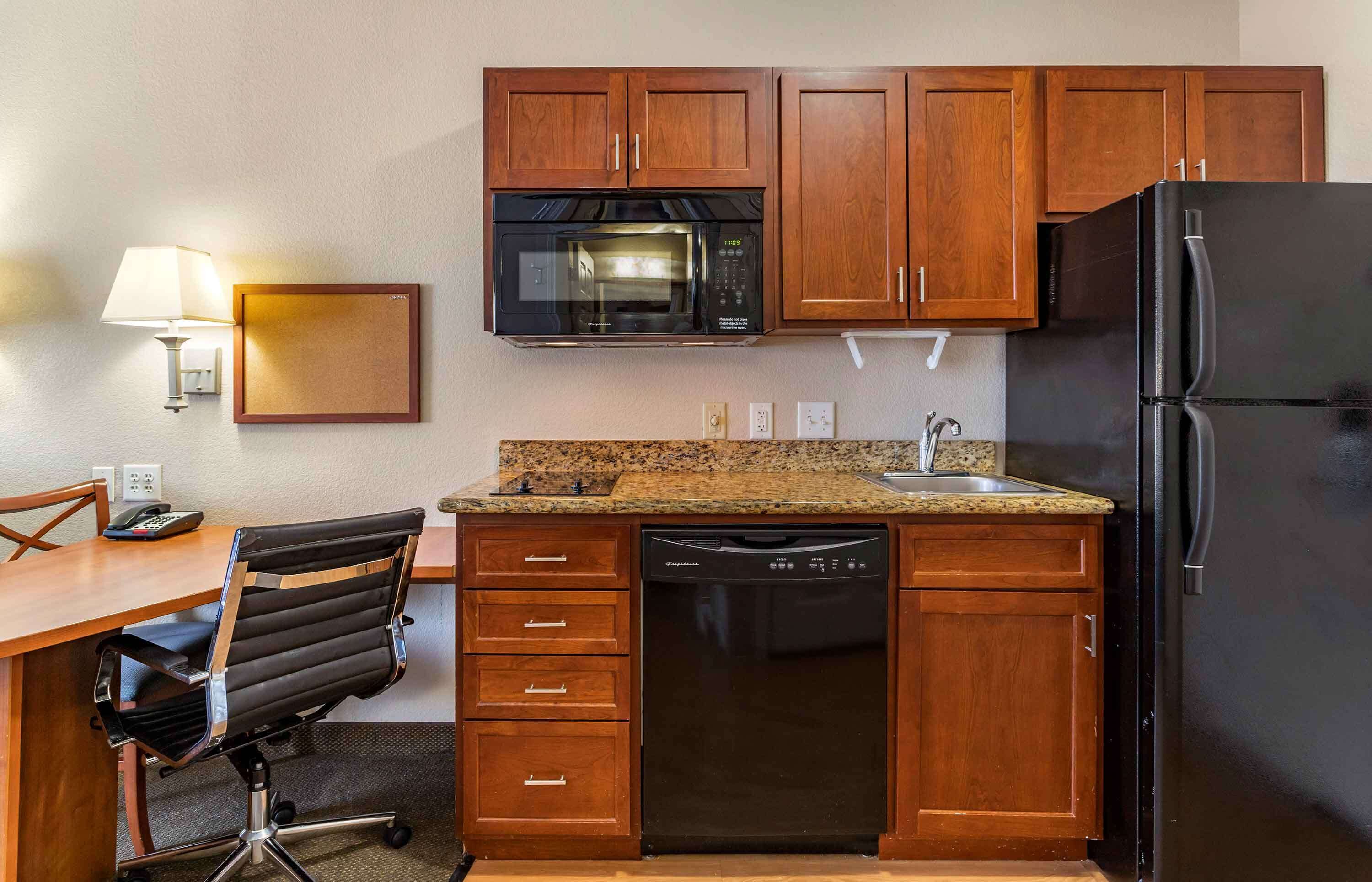 A compact kitchen with wooden cabinets, a microwave, a refrigerator, a dishwasher, and a desk with a chair.