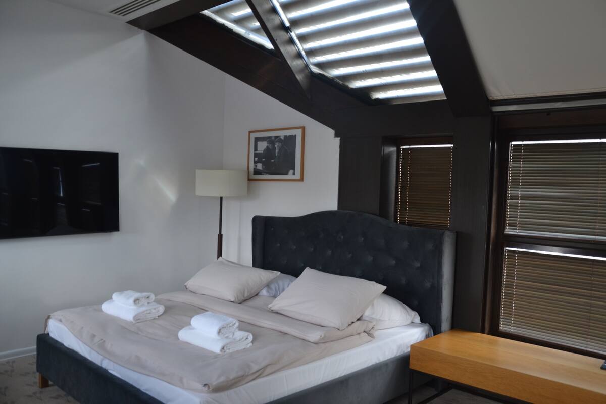 A neatly made bed with pillows and folded towels, a bedside table, a wall-mounted TV, and a framed picture on the wall.