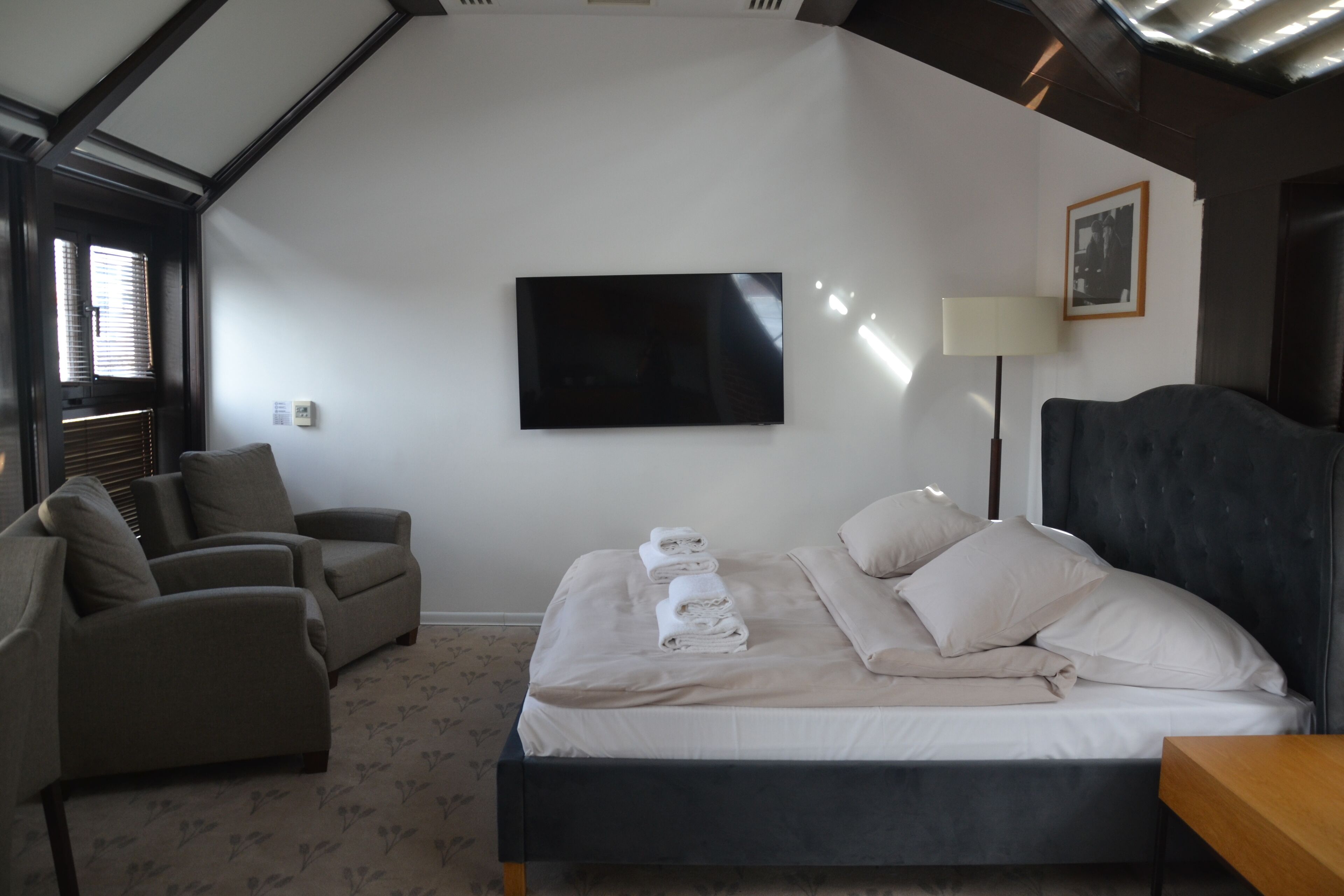 Un dormitorio con una cama, dos sillones, un televisor de pantalla plana y una lámpara de noche.