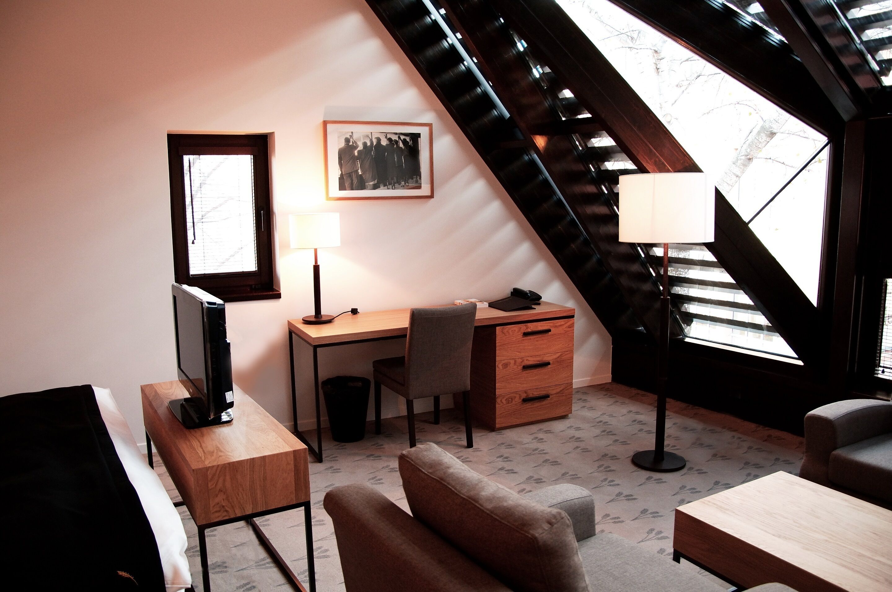 A modern room with a desk, chair, and a television. There is a window with a view of trees and a staircase.