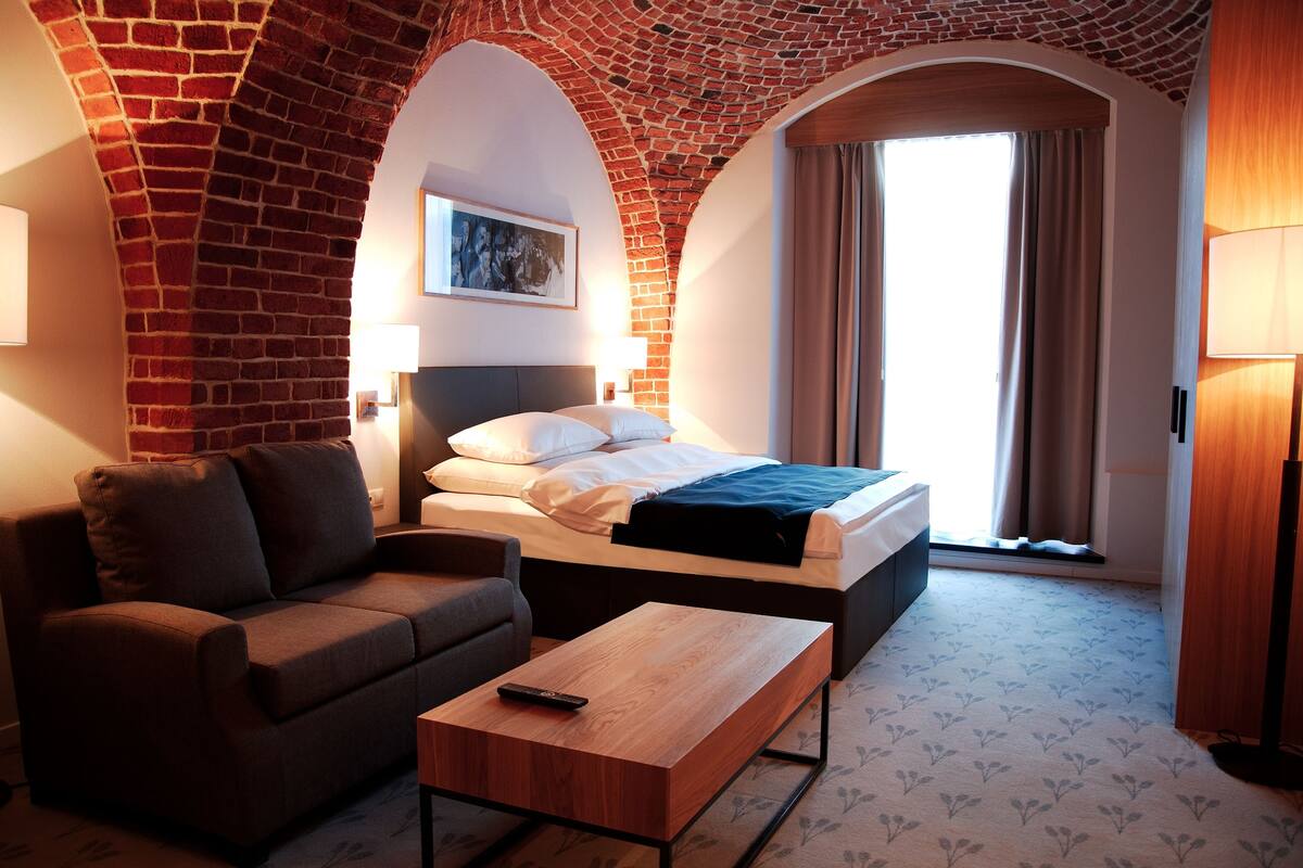 A hotel room with a brick arch, a bed, a sofa, a coffee table, and a lamp.